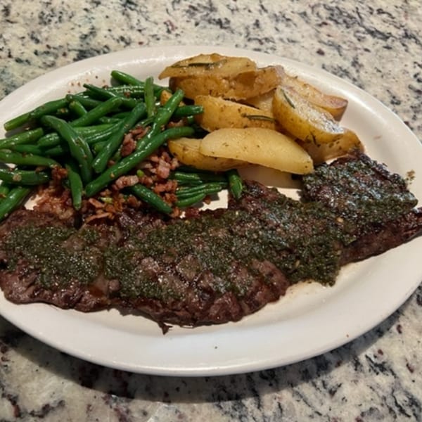 Grilled steak with green chimichurri sauce, roasted potato wedges, and green beans with bacon bits.