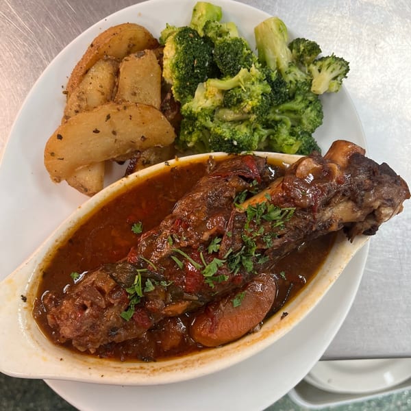 Braised lamb shank with gravy, roasted potato wedges, and steamed broccoli on a white plate.