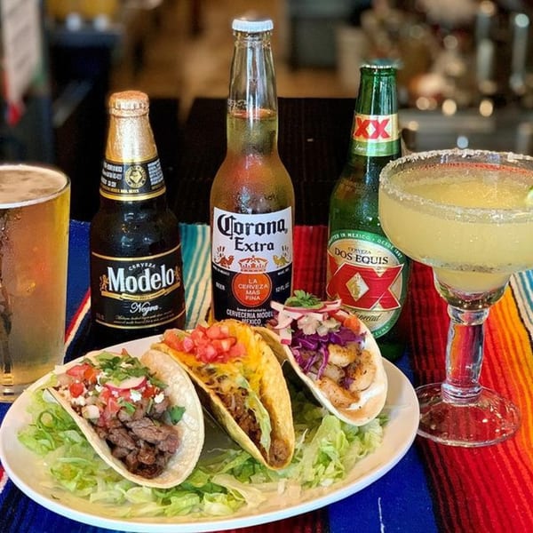 Three tacos, a margarita, and three bottles of Mexican beer on a colorful striped tablecloth.
